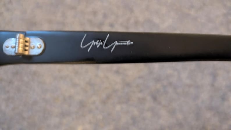 May include: Close-up of a black eyeglass arm with a silver hinge and the designer's signature in silver. The arm is rectangular and has a matte finish.
