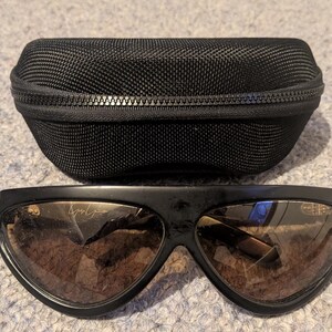 May include: Black sunglasses with brown lenses and a matching black case. The sunglasses have a wide, curved frame and a subtle logo on the lens. The case is zippered and made of a textured material, providing protection for the eyewear.