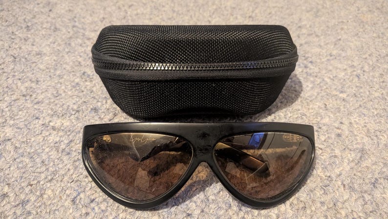 May include: Black sunglasses with brown lenses and a matching black case. The sunglasses have a wide, curved frame and a subtle logo on the lens. The case is zippered and made of a textured material, providing protection for the eyewear.