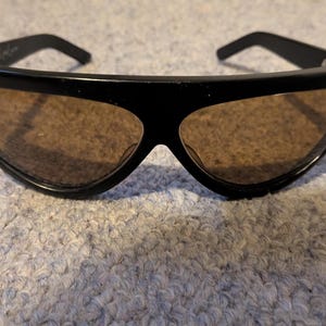 May include: Black sunglasses with brown tinted lenses. The sunglasses have a thick, curved frame and a sleek, modern design. The lenses are a warm brown color, and the frame is a glossy black.