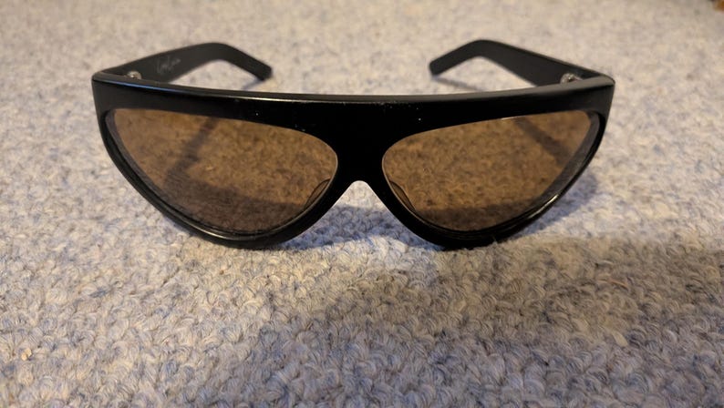 May include: Black sunglasses with brown tinted lenses. The sunglasses have a thick, curved frame and a sleek, modern design. The lenses are a warm brown color, and the frame is a glossy black.