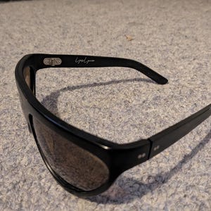 May include: Black sunglasses with a wide frame and tinted lenses. The sunglasses have a sleek, modern design with the text "Lyndia" on the arm. The sunglasses are suitable for outdoor activities and offer protection from the sun.