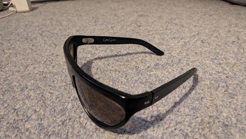 May include: Black sunglasses with a wide frame and tinted lenses. The sunglasses have a sleek, modern design with the text "Lyndia" on the arm. The sunglasses are suitable for outdoor activities and offer protection from the sun.