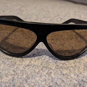 May include: Black sunglasses with a thick, angular frame and brown-tinted lenses. The sunglasses have a retro design, with a wide top and a distinctive shape. The lenses are a warm brown color, and the frame is a solid black.