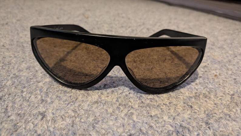 May include: Black sunglasses with a thick, angular frame and brown-tinted lenses. The sunglasses have a retro design, with a wide top and a distinctive shape. The lenses are a warm brown color, and the frame is a solid black.