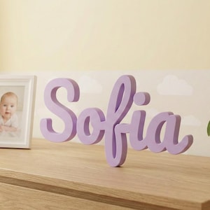 Puede incluir: Una cómoda de madera con una foto de bebé enmarcada, bloques de madera y un letrero de nombre de madera de color lavanda que dice "Sofia". Una pequeña planta en maceta con hojas verdes está a la derecha.