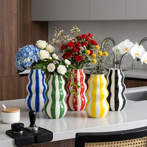 May include: A collection of five ceramic vases with wavy silhouettes, each adorned with vertical stripes in various colors: blue, green, red, yellow, and black. The vases hold an assortment of fresh flowers, including hydrangeas, roses, and orchids.