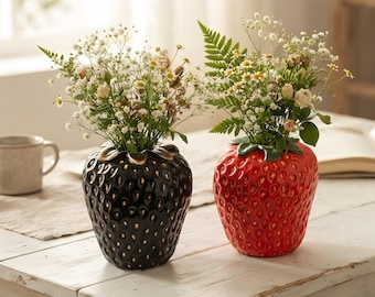 May include: Two ceramic strawberry-shaped vases, one black and one red, each featuring a textured surface with small indentations and green leaves at the top. Both vases are filled with a bouquet of small white flowers and green fern leaves.