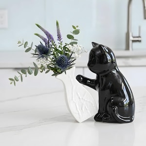 Puede incluir: Una figura de gato de cerámica negra brillante, interactuando con un jarrón blanco que contiene flores. El jarrón tiene un diseño de hojas en relieve. Las flores incluyen cardos azules y flores moradas.
