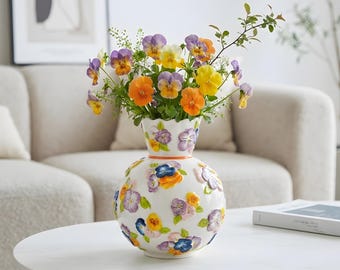May include: A white ceramic vase with a spherical body and a narrow neck, decorated with raised, three-dimensional painted pansy flowers in shades of purple, yellow, and orange. The vase is filled with a bouquet of fresh, colorful pansies.