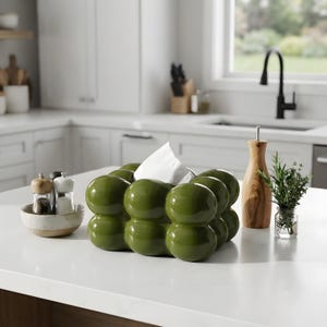 May include: A glossy olive green tissue box cover featuring a rounded, bubble-like exterior design. The rectangular container has a central opening at the top from which a single white tissue is pulled upward, showcasing a smooth, reflective finish.