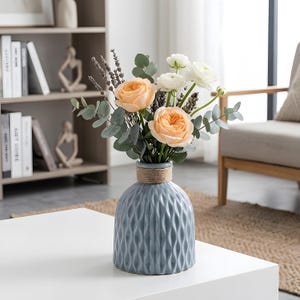 May include: A blue ceramic vase with a textured, diamond-patterned surface, holding a bouquet of peach roses, white flowers, and greenery. The vase has a rope detail around the neck and sits on a white table.