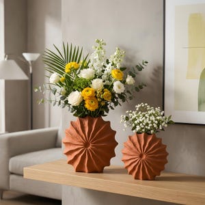 May include: Two terracotta-colored vases with a sunburst design hold fresh flowers. The larger vase contains yellow, white, and green flowers, while the smaller vase holds white baby's breath. The vases sit on a wooden shelf.