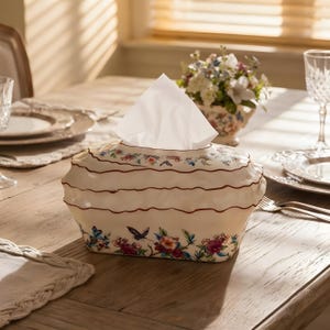 May include: A cream-coloured ceramic tissue box cover with a wavy, tiered silhouette and a brown-painted rim. The base features a colourful floral and butterfly pattern, while a single white tissue extends from the top opening of the container.