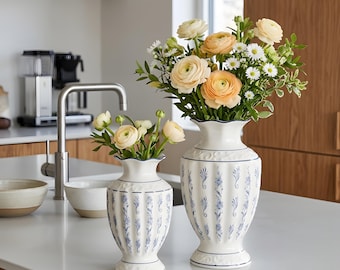 May include: Two white ceramic vases with blue floral patterns and ruffled rims, filled with fresh flowers. The vases are of different sizes and sit on a white countertop, showcasing a kitchen setting.