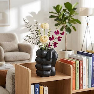 May include: A matte black ceramic vase shaped like a clenched fist, holding a bouquet of two cream-colored ranunculus flowers, several sprigs of green eucalyptus, and a few stems of deep magenta sweet pea flowers, resting on a wooden shelf.