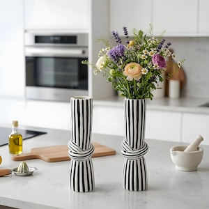 Könnte beinhalten: Zwei hohe zylindrische Keramikvasen mit einem schwarz-weißen vertikalen Streifenmuster und einem dekorativen Knotendetail in der Mitte. Eine Vase ist leer, während die andere einen bunten Strauß aus Lavendel, Gänseblümchen und Ranunkeln enthält.