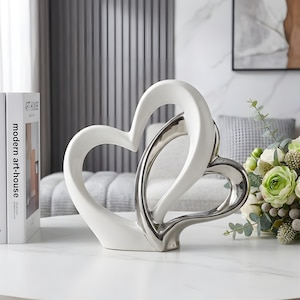 May include: A decorative sculpture featuring two interlocking heart shapes. The larger outer heart is finished in a smooth, glossy white, while the smaller inner heart has a reflective, metallic silver finish. The piece stands on a flat base.
