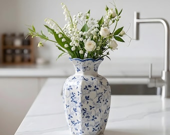 May include: A white ceramic vase with a scalloped rim and a blue floral pattern, featuring a hexagonal body shape. The vase is filled with a bouquet of white roses and lily of the valley flowers with green stems and leaves.