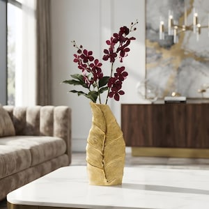 Op de afbeelding: Een goudkleurige vaas in de vorm van een groot, getextureerd blad met een prominente centrale nerf en horizontale ribbels. De vaas bevat verschillende stengels met diepbordeauxrode orchideeën en groene bladeren, wat een opvallend contrast vormt tussen het metallic goud en de donkere bloemtinten.