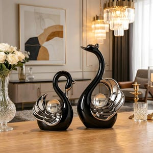 May include: Two swan figurines crafted from glossy black ceramic with silver-toned wing accents. Each swan features a clear, spherical glass orb integrated into the center of its body. The pair consists of one smaller and one larger swan sculpture.
