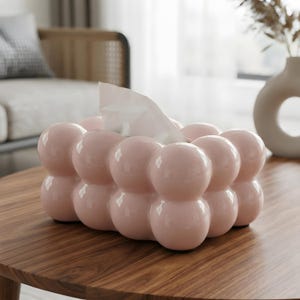 May include: A rectangular tissue box cover featuring a glossy, light pink finish with a distinct bubble-like, spherical texture across its exterior. The structure is composed of multiple rounded, ball-shaped segments, with a white tissue protruding.