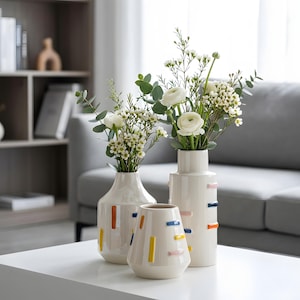 May include: A set of three off-white ceramic vases of varying heights, each decorated with small, raised rectangular accents in orange, blue, yellow, and pink. Two of the vases contain arrangements of white flowers and green eucalyptus leaves.
