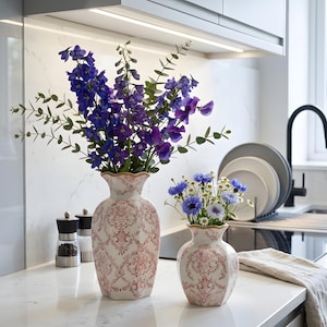 May include: Two decorative vases with floral arrangements. The larger vase holds purple flowers, while the smaller one contains blue and white flowers. Both vases have a white and red pattern with a gold rim. The vases are on a white countertop.
