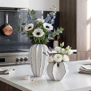 Könnte beinhalten: Zwei weiße Keramikvasen mit blauen Akzenten, gefüllt mit frischen Blumen, stehen auf einer weißen Arbeitsplatte. Die größere Vase hat vertikale blaue Streifen, während die kleinere einen wellenförmigen blauen Rand hat. Die Blumen umfassen weiße Anemonen, blaue Rittersporne und weiße Rosen.