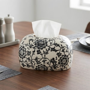 Puede incluir: Una caja rectangular de cerámica para pañuelos con una base de color crema y un patrón floral negro detallado. El diseño consiste en flores estilizadas repetitivas y enredaderas que cubren toda la superficie exterior del soporte.