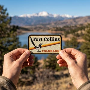 May include: A rectangular embroidered patch featuring a black and white magpie bird perched on a horizontal orange line. The text reads Fort Collins in black letters and Colorado in orange letters against a cream-colored background with a black border.