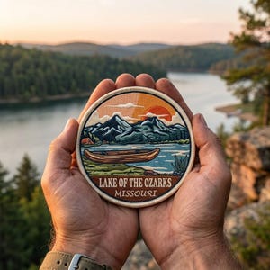 May include: A round patch featuring a scenic landscape with a lake, mountains, and a canoe. The patch has the text "Lake of the Ozarks Missouri" in white. The patch is held in two hands against a blurred background of a lake and trees.