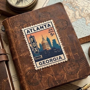 May include: A brown leather-bound journal featuring a rectangular postage stamp graphic on the cover. The stamp displays the text ATLANTA, AIR MAIL, 5c, FOX, and GEORGIA, with an illustration of a city skyline and a domed building at sunset.