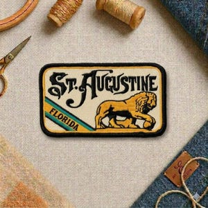 May include: A rectangular patch with the text "St. Augustine Florida" and a lion illustration. The patch has a black border, a cream-colored background, and yellow and teal accents. The lion is yellow and black.