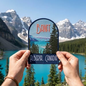 May include: A semi-circular embroidered patch featuring a landscape of Banff, Alberta, Canada. The design includes a light blue sky, a turquoise lake, and dark green pine trees. The text BANFF is in orange, and ALBERTA, CANADA is in white.