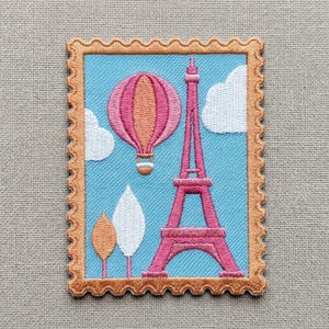 May include: A rectangular embroidered patch shaped like a postage stamp with a scalloped tan border. The design features a pink Eiffel Tower, a pink and orange hot air balloon, and white clouds against a light blue sky background.