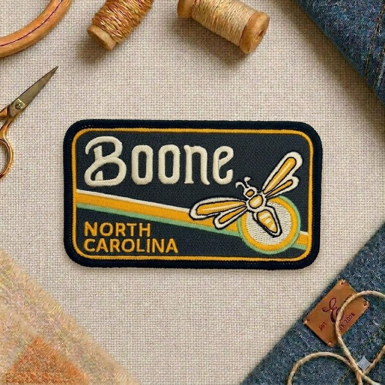 May include: A rectangular patch with the word "Boone" in white, arched text above "North Carolina". A yellow and white dragonfly design is on the right. The patch has a navy blue background with a yellow and green stripe.