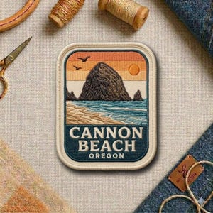 May include: A rectangular embroidered patch featuring a landscape of Cannon Beach, Oregon, with a large dark rock formation, blue ocean waves, a sandy beach, and a sunset sky with two birds. The text CANNON BEACH OREGON is stitched in white.