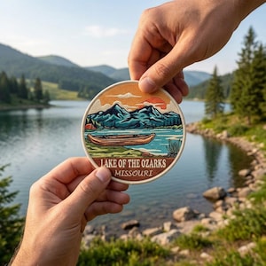 May include: A round patch featuring a scenic lake landscape with mountains, a canoe, and a sunset. The text "Lake of the Ozarks Missouri" is displayed at the bottom. The patch is held up against a lake background.