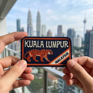 May include: A rectangular embroidered patch with a dark blue background, featuring the text KUALA LUMPUR in white, an orange and black tiger illustration, and a diagonal orange stripe with the word MALAYSIA in black text on a white background.