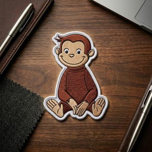 May include: Embroidered patch featuring a seated cartoon monkey with a brown body, tan face, and a white border. The monkey is smiling with its hands clasped in front. The patch is on a wooden surface.