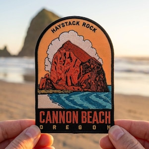 May include: An embroidered patch featuring a landscape of Haystack Rock at Cannon Beach, Oregon. The design includes a large orange rock formation, white clouds, and blue ocean waves. The text reads HAYSTACK ROCK, CANNON BEACH, OREGON.