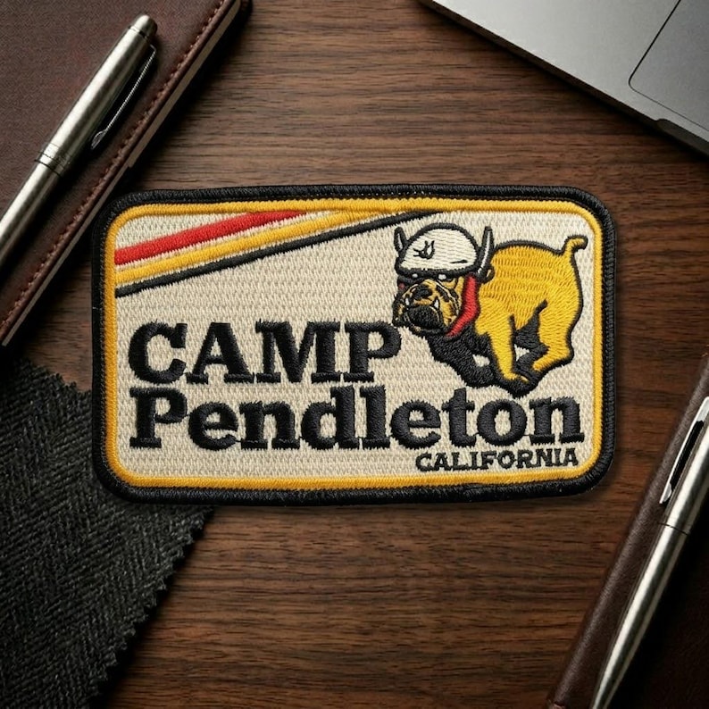 May include: Embroidered patch featuring the text "CAMP Pendleton CALIFORNIA" with a yellow bulldog wearing a helmet. The patch has a cream background with red and yellow stripes and a black border.