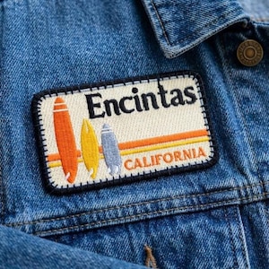 May include: Embroidered patch featuring the word "Encintas" above three surfboards in orange, yellow, and blue. Below the surfboards are orange and yellow stripes and the word "CALIFORNIA". The patch is on a blue denim jacket.