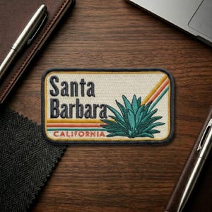 May include: Embroidered patch featuring the text "Santa Barbara CALIFORNIA" with a teal agave plant design and retro stripes. The patch has a cream-colored background and a black border. A travel-themed souvenir.