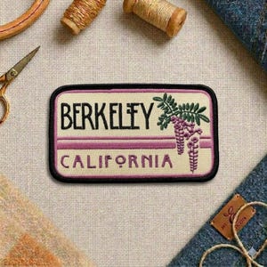 May include: Embroidered patch featuring the city name "Berkeley" above "California" in black text. The patch has a cream background with purple stripes and a floral design. The patch is rectangular with a black border.