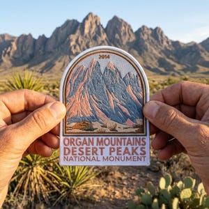 May include: A patch featuring a mountain range design with the text "ORGAN MOUNTAINS DESERT PEAKS NATIONAL MONUMENT" and the year "2014". The patch has a white border and is held up in front of a desert landscape.