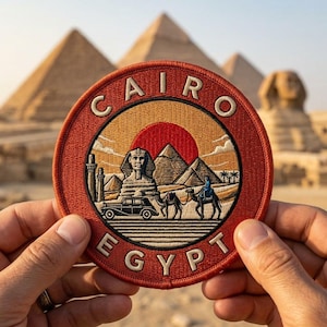 Peut inclure: Un écusson brodé circulaire avec une bordure rouge portant le texte CAIRO EGYPT. Le centre représente un soleil rouge derrière le Sphinx et les pyramides, avec une voiture ancienne et une personne à dos de chameau au premier plan, dans des tons chauds.