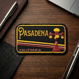 May include: Embroidered patch featuring the word "Pasadena" in yellow script above a stylized image of a street lamp. The patch has a black background with yellow and red horizontal stripes and the word "CALIFORNIA" below.