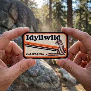 May include: A rectangular embroidered patch with a cream-colored background and a dark blue border. The text Idyllwild appears in dark blue, with California below. An illustration of a brown chipmunk sits beside three diagonal orange and brown stripes.